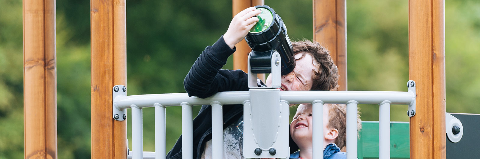 Young children interact with a telescope at a playground, they are peeking through and looking up at the sky.